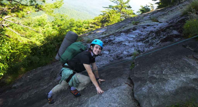 a person wearing rock climbing gear smiles at the camera as they scale a rock on an outward bound trip for lgbtq teens